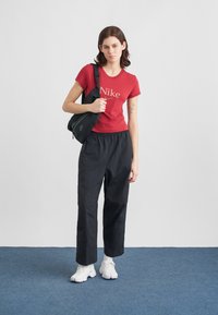 Νεαρή γυναίκα φορώντας κόκκινο μπλουζάκι Nike, μαύρο παντελόνι, λευκά παπούτσια και κρατώντας μια μαύρη τσάντα ώμου σε απλό φόντο.