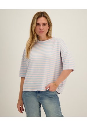 Vrouw met lang blond haar, gekleed in een losse roze en lichtblauw gestreepte blouse en lichtblauwe jeans, poserend met één hand in de zak.