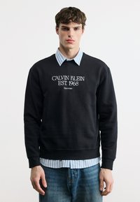 Νεαρός άνδρας που φοράει μαύρο φούτερ Calvin Klein με το κείμενο "EST. 1968" πάνω από ριγέ πουκάμισο με κουμπιά και μπλε τζιν, στέκεται μπροστά σε λευκό φόντο.