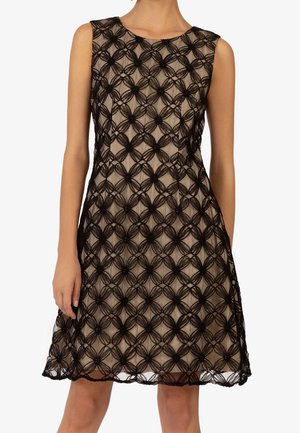 Ärmelloses beiges Kleid mit schwarzer Spitzenüberlagerung mit Blumenmuster, getragen von einer stehenden Person, knielang mit rundem Ausschnitt.