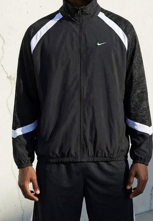 Homme portant une veste de survêtement Nike noire et blanche avec fermeture éclair intégrale à l'avant et taille élastique, debout devant un mur clair.