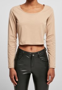 Beige Langarm-Crop-Top aus weichem Stoff, mit Rundhalsausschnitt. Kombiniert mit hochgeschnittenen schwarzen Hosen aus Kunstleder mit Taschen.