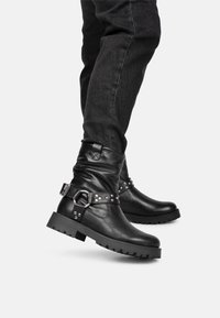 Botas de cuero negras hasta el tobillo con un diseño caído, que cuentan con tachuelas metálicas y un detalle de hebilla. Suela de goma gruesa para mayor durabilidad.