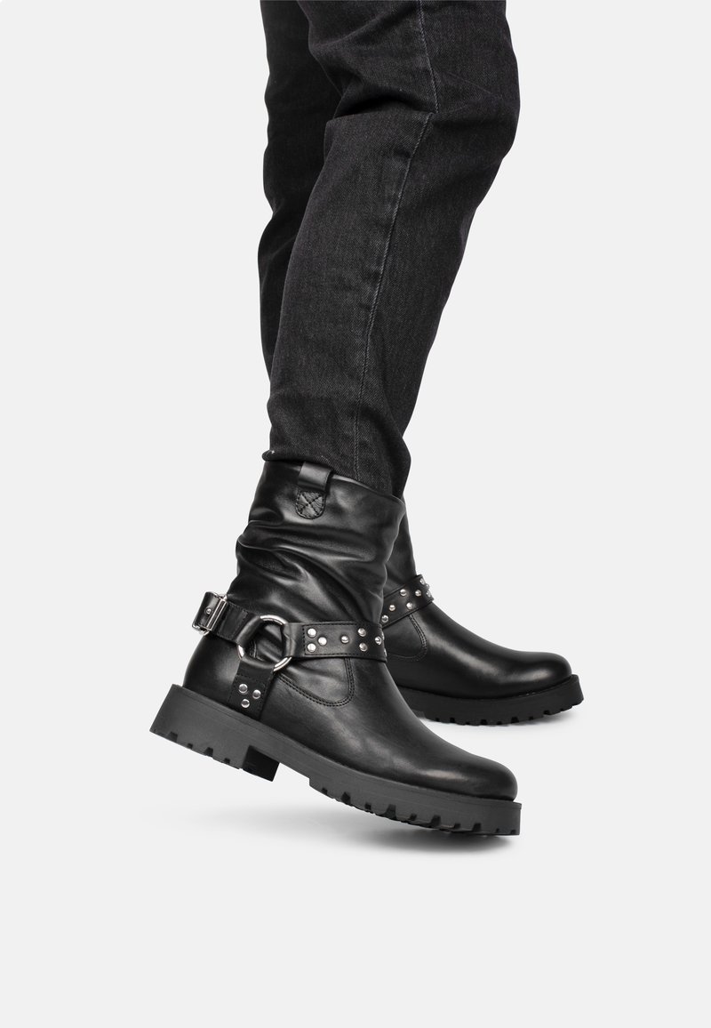 Botas de cuero negras hasta el tobillo con un diseño caído, que cuentan con tachuelas metálicas y un detalle de hebilla. Suela de goma gruesa para mayor durabilidad.