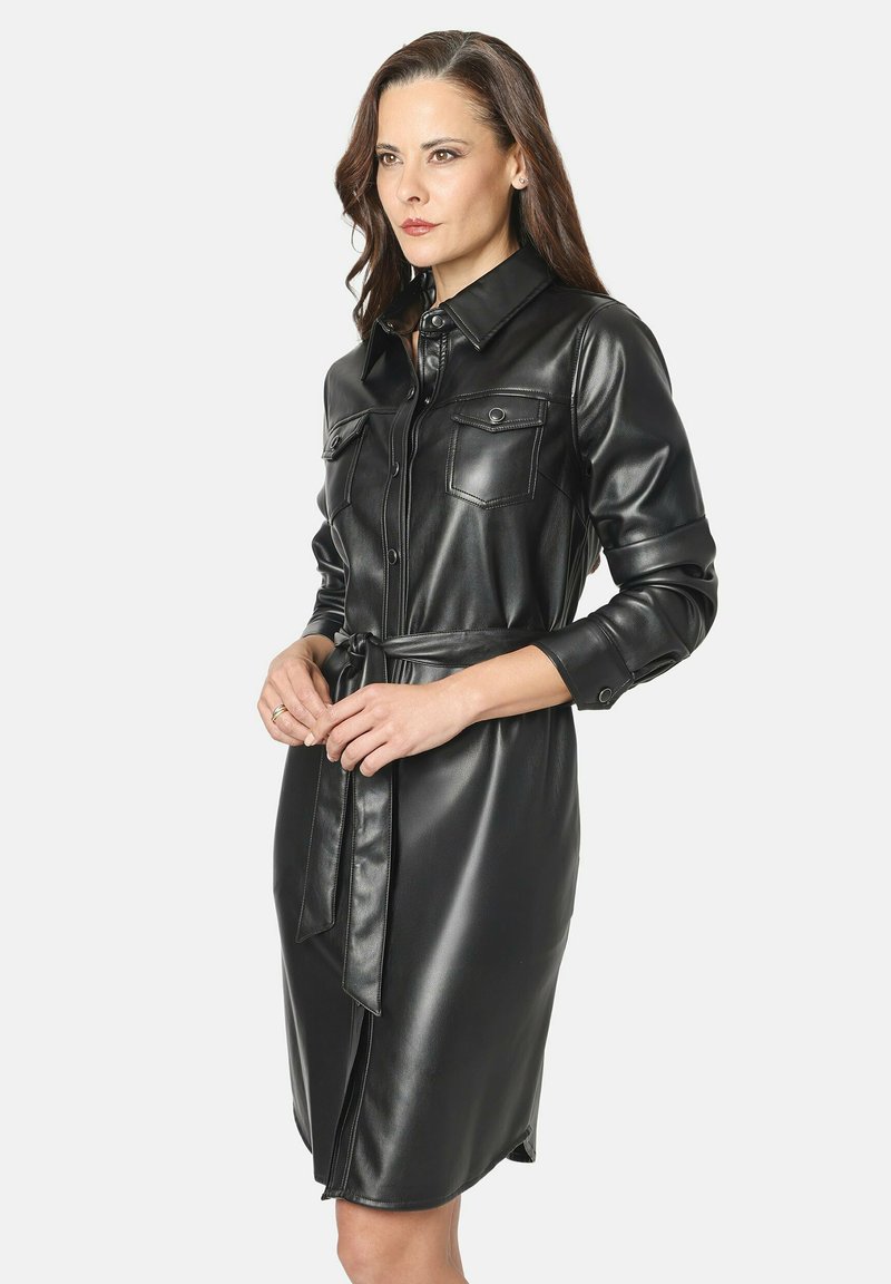 Mujer con cabello castaño de longitud media que lleva un vestido camisero negro de piel sintética con cinturón, de mangas largas y con bolsillos en el pecho abotonados.