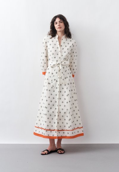 Femme portant une longue robe blanche avec de petits motifs floraux noirs, bordure orange aux poignets et à l'ourlet, taille nouée, debout devant un mur blanc uni.