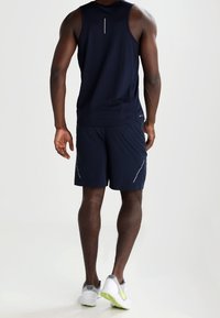 Débardeur de sport bleu marine à coupe ajustée, design sans coutures et accents réfléchissants ; porté avec un short assorti et des baskets blanches.