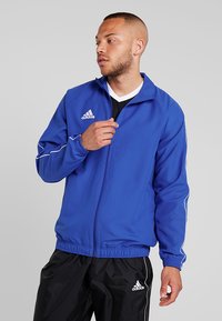Leichte blaue Jacke mit Stehkragen, ausgestattet mit weißen Paspeln, Reißverschluss und dem Adidas-Logo auf der Brust und dem Arm.
