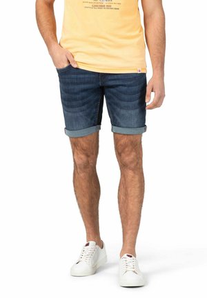 Dunkelblaue Denim-Shorts mit hochgerollten Säumen, die eine verblasste Textur und seitliche Taschen besitzen. Kombiniert mit weißen Turnschuhen und einem gelben T-Shirt mit Grafik.