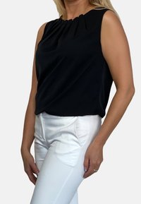 Musta hihaton paita, jossa on laskostettu kaula-aukko, yhdistettynä tiukkoihin valkoisiin housuihin, jotka korostavat sileää tekstuuria ja kontrastivärejä.