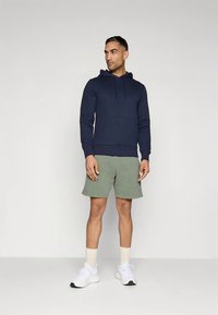 Temno modra bombažna jopa z žepom spredaj in vrvicami za zategovanje, usklajena z zelenimi bombažnimi kratkimi hlačami. Bele superge in svetlo obarvane nogavice običajne dolžine.