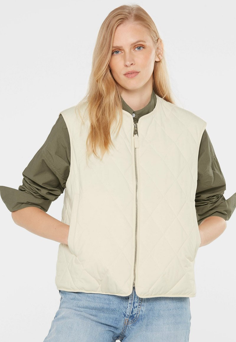 Junge Frau mit langen blonden Haaren, die eine cremefarbene gesteppte Weste über einem olivgrünen Hemd und hellblauen Jeans trägt, steht mit den Händen in den Taschen.