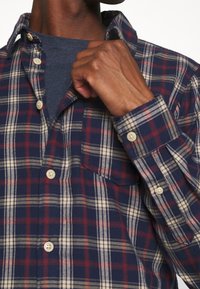 Camicia a flanella a quadri in blu navy, rosso e crema. Presenta una chiusura con bottoni, taschino sul petto e maniche arrotolate. Tessuto morbido.