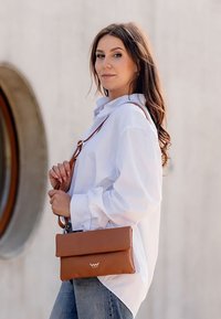 Hnědá kožená taška přes tělo s překlopným designem, s hladkým povrchem a stříbrným logem. Nosená s bílou košilí a džíny.