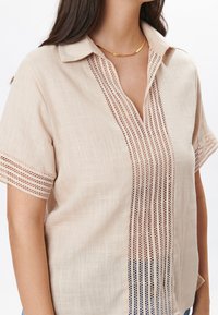 Blusa corta de manga corta en beige claro con cuello tipo camisa, que presenta paneles verticales de encaje de ojal y una textura sutil.