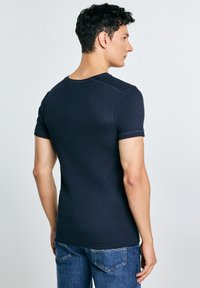 Mand med kort krøllet hår iført en tætsiddende marineblå kortærmet skjorte og blå jeans, stående og vendt sidelæns mod en ensfarvet baggrund.