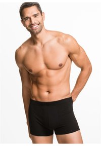 Boxer corti neri in cotone con un elastico indossati da un uomo con un fisico tonico. Sfondo bianco semplice.