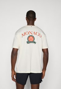 Kremowa koszulka z krótkim rękawem z dużym czerwonym napisem "MONACO" oraz okrągłym zielonym logo z białymi akcentami. Gładka faktura materiału.