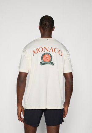 Krémové tričko s krátkym rukávom s veľkým červeným nápisom "MONACO" a kruhovým zeleným logom s bielymi akcentmi. Hladká textúra látky.