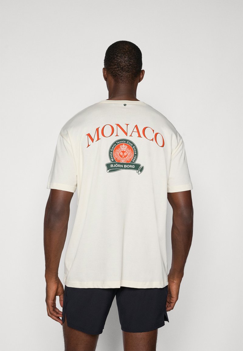 Kremowa koszulka z krótkim rękawem z dużym czerwonym napisem "MONACO" oraz okrągłym zielonym logo z białymi akcentami. Gładka faktura materiału.