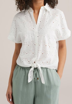 Kobieta w białej krótkiej koszuli zapinanej na guziki z szyfonową koronką, związanej na talii, oraz w jasnoszarych spodniach ze sznurkiem.