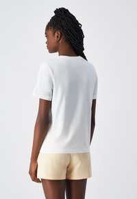 Weißes Baumwoll-T-Shirt mit Rundhalsausschnitt und kurzen Ärmeln, von hinten gezeigt. Lockerer Schnitt mit glatter Textur, kombiniert mit hellbeigen Shorts.