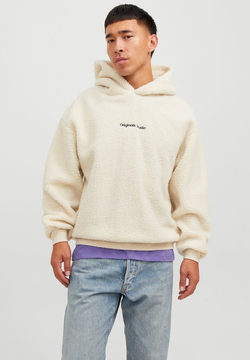 Kremfarget hettegenser laget av teksturert fleece, med et lappemerke, ribbede mansjetter, og lagd over en lilla skjorte.