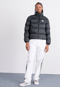 Zwarte teddy jas met een hoge kraag en rits, voorzien van een klein wit logo. Gecombineerd met witte broek met zwarte zijstrepingen en grijze sneakers.