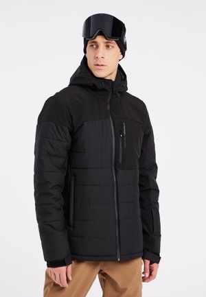 Schwarze isolierte Jacke mit einer Reißverschlusstasche auf der Brust, gestepptem Design, hohem Kragen und verstellbare Kapuze. Kombiniert mit beigen Hosen.