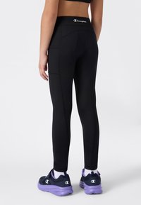Personne portant un legging noir Champion avec une poche latérale et des baskets navy avec des semelles violettes, se tenant contre un fond uni.