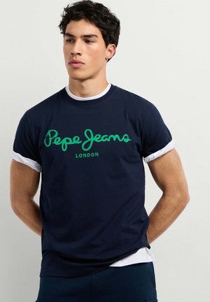 Giovane uomo con capelli scuri che indossa una maglietta blu navy Pepe Jeans London sopra una camicia bianca a maniche lunghe, in piedi con le mani dietro la schiena.