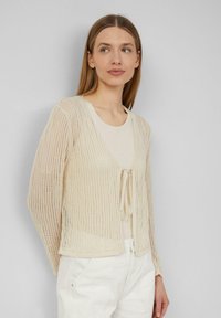 Beige, offenes Strickcardigan mit langen Ärmeln, versehen mit einer Bindeschließe vorne. Getragen über einem hellen Oberteil, mit weicher Textur.