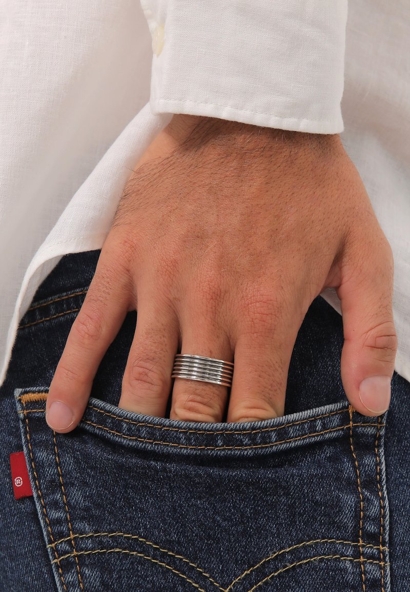 Metallring med växelvis släta och texturerade band, bärs på en hand som vilar i en denimficka, med en vit skjorta delvis synlig.