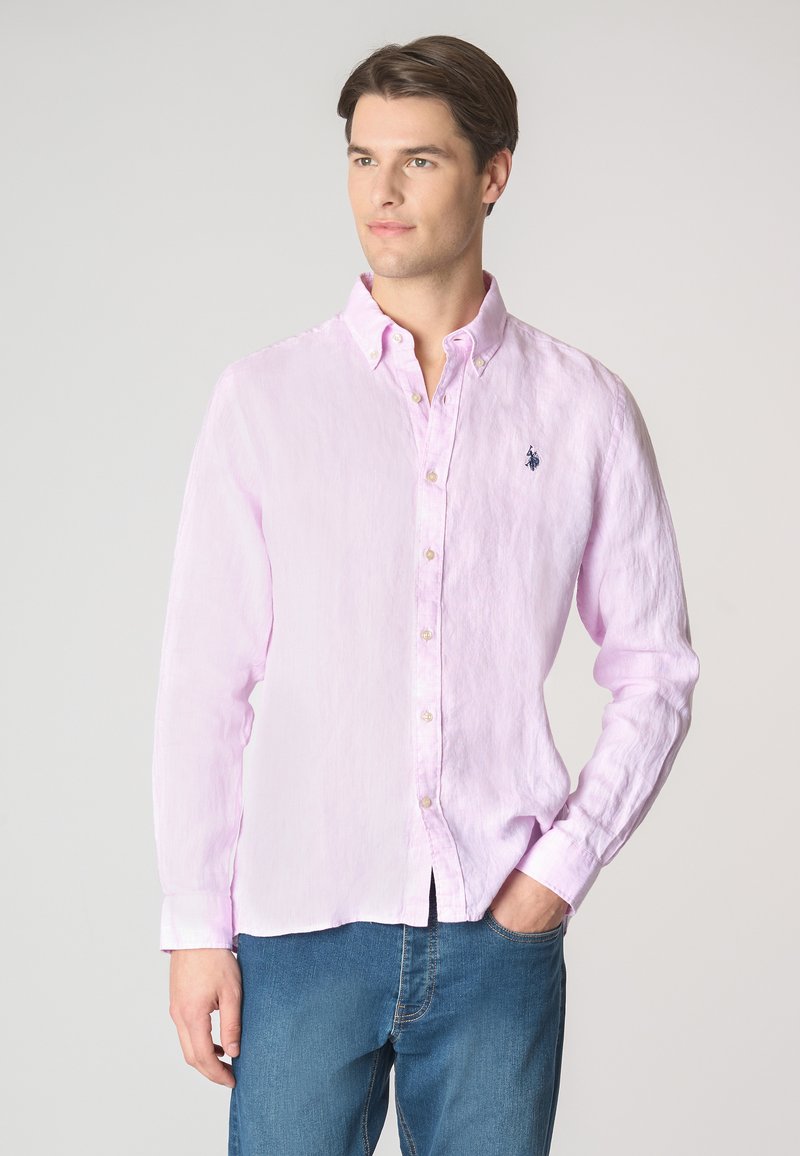 Giovane uomo con capelli corti e ben curati, indossa una camicia button-up rosa chiaro e jeans blu, in piedi contro uno sfondo semplice, sorridendo delicatamente.