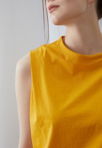 Sleeveless yellow cotton top with a round neckline, featuring smooth texture and minimal stitching details, set against a light background.