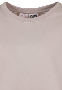 Lys beige bomulls T-skjorte med rund halslinning. Har en etikett med teksten "URBAN CLASSICS" og pleieinstruksjoner nær kragen. Glatt tekstur.