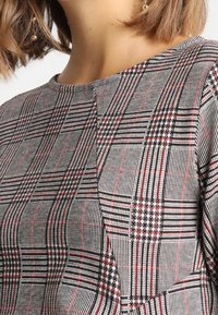 Tissu à motif à carreaux gris et noirs avec des accents rouges, présentant un col rond et une texture finie.