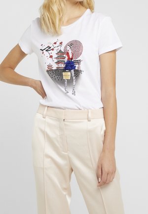 Kobieta w białej koszulce z kolorową grafiką Tokio, beżowych spodniach, ręka na biodrze, blond włosy, neutralne szare tło.