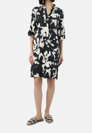 Vrouw draagt knielange zwart-witte bloemenjurk met opgerolde driekwart mouwen en instapsandalen tegen een effen achtergrond.