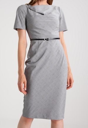 Frau trägt ein figurbetontes, knielanges graues kariertes Kleid mit kurzen Ärmeln und einem schwarzen Gürtel in der Taille vor einem einfarbigen Hintergrund.