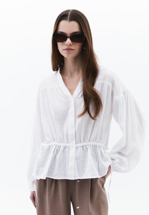 Chemisier blanc à boutons avec une taille peplum, de longues manches bouffantes et un tissu texturé. Associé à un pantalon marron et des lunettes de soleil surdimensionnées.