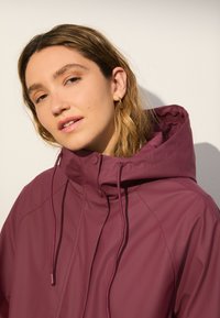 Vínovočervená nepremokavá bunda s vysokým golierom, sťahovacími šnúrkami, patentnými gombíkmi a hladkou textúrou. Dizajn je funkčný a priliehavý.