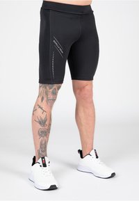 Zwarte sportshorts van Lycra, met een gladde textuur, reflecterend logo en stippenaccenten, gecombineerd met witte hardloopschoenen.