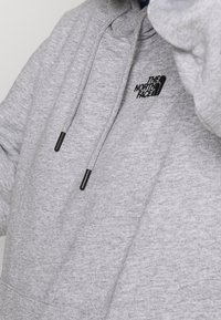 Šedá bavlněná mikina s kapucí na šňůrku, s černým logem na hrudi a jemnou texturou. Žádné viditelné vzory ani akcenty.