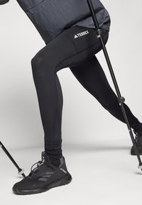 Sorte strækleggings med Terrex-logo, der har flade sømme og en tætsiddende pasform. Begge dele ledsaget af sorte vandresko og en vandrestav.