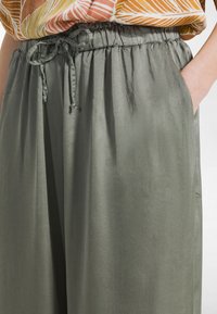 Olivengrønne, lette bukser i satinstof med elastisk talje og snøre. Sidelommer synlige, glat tekstur.