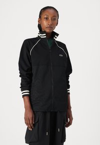 Žena s pletenicom koja nosi crnu jaknu na zip s bijelim trakama na ovratniku i rukavima, stoji naslonjena na jednostavnu bijelu pozadinu.