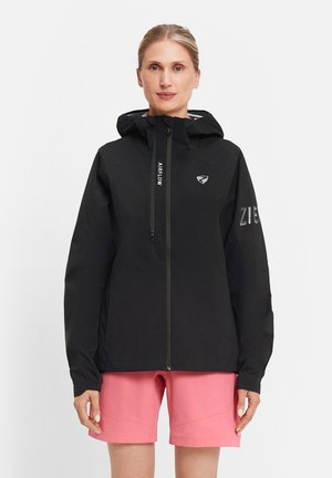 Frau trägt schwarze Kapuzenjacke mit Reißverschluss und dem Schriftzug "Airflow" sowie pinke Shorts, steht vor einem schlichten weißen Hintergrund.