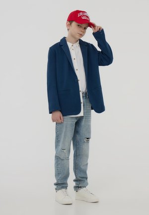 Blazer blu con rever, camicia bianca a maniche lunghe, jeans chiari strappati, cappellino rosso con la scritta "Los Angeles California" e sneaker bianche.