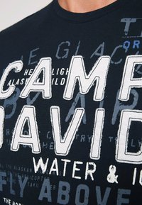 Czarna bawełniana koszulka z wyraźnymi białymi i szarymi wzorami tekstowymi, w tym "CAMP DAVID" oraz różnymi grafikami, z okrągłym dekoltem.